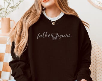 May include: A black sweatshirt with the words "father figure" in cursive script. A football helmet graphic is next to the word "figure". The sweatshirt is a dark color and has long sleeves.