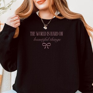 Może przedstawiać: Czarna bluza z napisem "THE WORLD IS HARD ON beautiful things" w jasnor&oacute;żowym kolorze, wraz z graficznym łukiem. Bluza ma okrągły dekolt i długie rękawy.