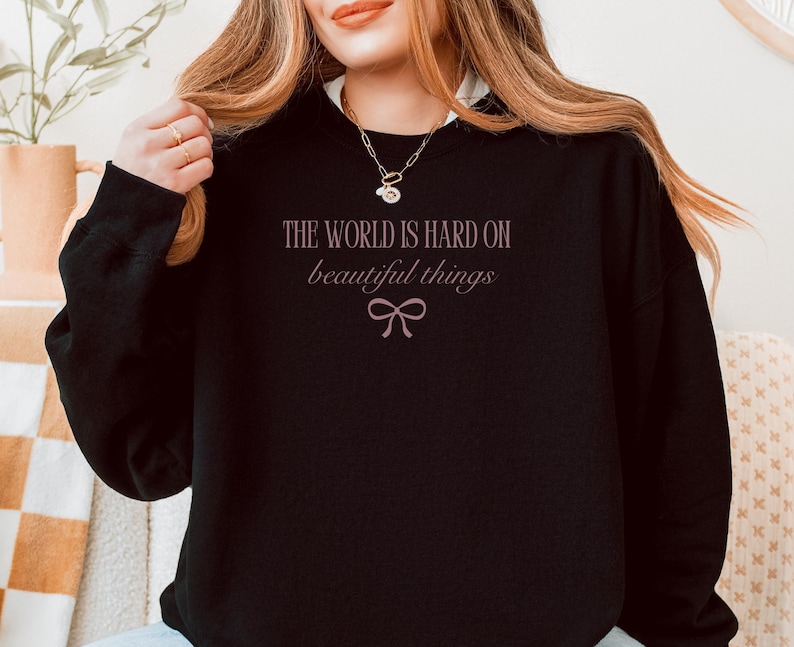 Może przedstawiać: Czarna bluza z napisem "THE WORLD IS HARD ON beautiful things" w jasnor&oacute;żowym kolorze, wraz z graficznym łukiem. Bluza ma okrągły dekolt i długie rękawy.