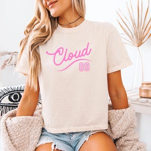 Puede incluir: Camiseta color crema con el texto "Cloud 09" en escritura rosa y letras de bloque. La camiseta es de corte holgado y tiene mangas cortas. La modelo lleva pantalones cortos vaqueros azules.