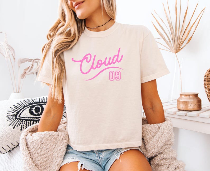 Puede incluir: Camiseta color crema con el texto "Cloud 09" en escritura rosa y letras de bloque. La camiseta es de corte holgado y tiene mangas cortas. La modelo lleva pantalones cortos vaqueros azules.