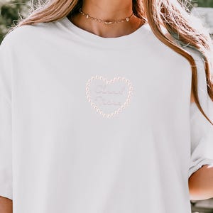 May include: White t-shirt with a heart-shaped design. The heart is outlined with small, light pink beads and contains the words "Cloud Nine" in a cursive font. The shirt has short sleeves and a crew neck.