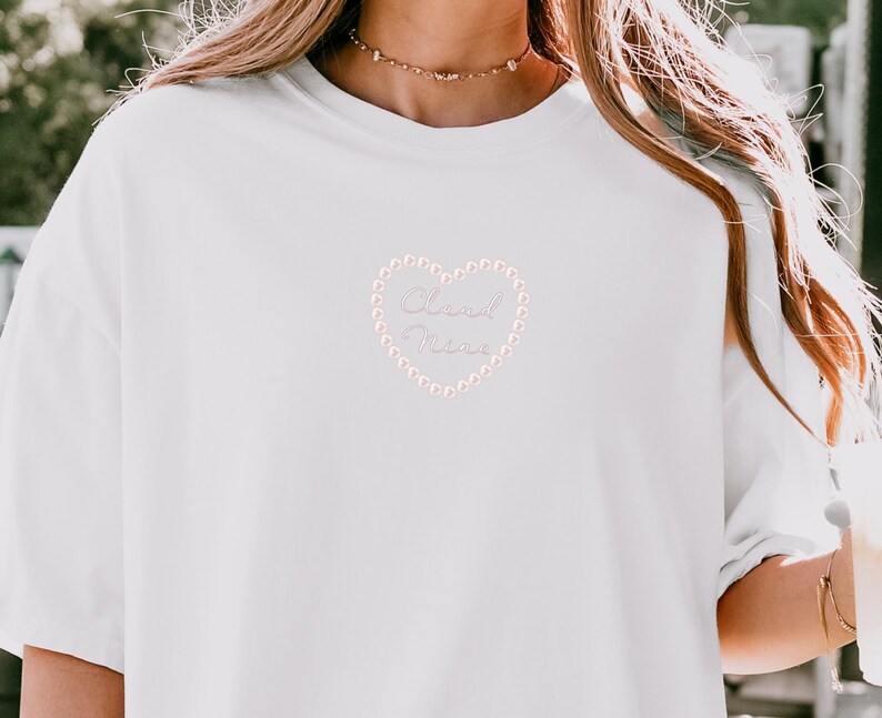 May include: White t-shirt with a heart-shaped design. The heart is outlined with small, light pink beads and contains the words "Cloud Nine" in a cursive font. The shirt has short sleeves and a crew neck.