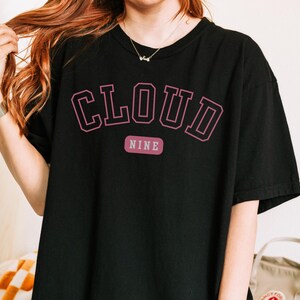 May include: Black t-shirt with the text "CLOUD NINE" in a pink, collegiate-style font. The word "NINE" is on a separate, smaller pink rectangle below the word "CLOUD". The shirt is a casual, oversized fit.