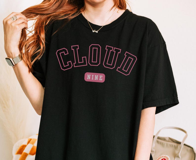 May include: Black t-shirt with the text "CLOUD NINE" in a pink, collegiate-style font. The word "NINE" is on a separate, smaller pink rectangle below the word "CLOUD". The shirt is a casual, oversized fit.