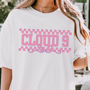 Puede incluir: Camiseta blanca con el texto "CLOUD 9" en rosa, enmarcado por un patr&oacute;n de cuadros rosas y morados. Un dise&ntilde;o de lazo rosa y morado est&aacute; debajo del texto. La camiseta es de estilo casual y de gran tama&ntilde;o.