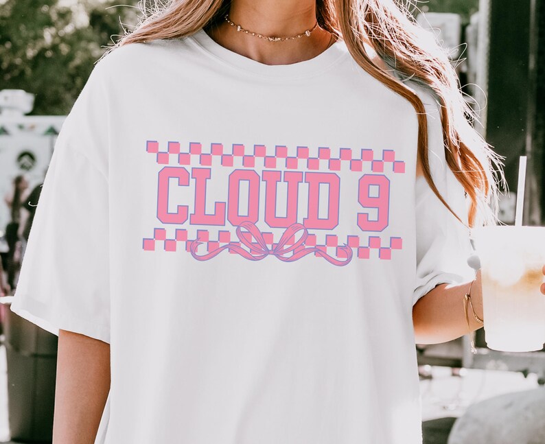 Puede incluir: Camiseta blanca con el texto "CLOUD 9" en rosa, enmarcado por un patr&oacute;n de cuadros rosas y morados. Un dise&ntilde;o de lazo rosa y morado est&aacute; debajo del texto. La camiseta es de estilo casual y de gran tama&ntilde;o.