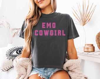 May include: Dark gray t-shirt with the text "EMO COWGIRL" in pink block letters. The shirt is a relaxed fit and appears to be made of a soft material. The model is wearing denim shorts and a cream-colored sweater.