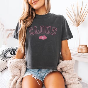May include: Dark gray t-shirt with the word "CLOUD" in pink block letters and a pink cloud graphic with the number 9. The shirt is a crew neck and has short sleeves. The model is wearing denim shorts and a cream-colored sweater.