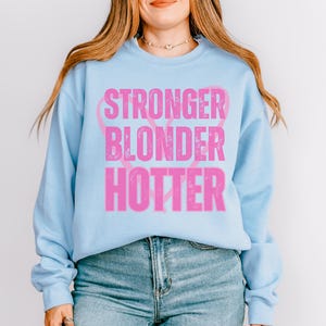 Puede incluir: Sudadera azul claro con las palabras "STRONGER BLONDER HOTTER" en letras rosas desgastadas. La sudadera tiene mangas largas y cuello redondo. El texto est&aacute; dispuesto verticalmente con un dise&ntilde;o de cinta rosa.