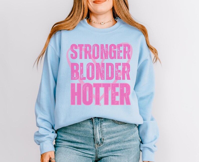 Puede incluir: Sudadera azul claro con las palabras "STRONGER BLONDER HOTTER" en letras rosas desgastadas. La sudadera tiene mangas largas y cuello redondo. El texto est&aacute; dispuesto verticalmente con un dise&ntilde;o de cinta rosa.