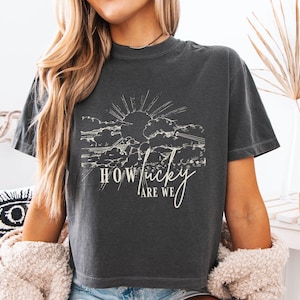 May include: Dark gray t-shirt with a white graphic of a sun rising over clouds. The text "HOW LUCKY ARE WE" is written in a cursive font below the graphic. The shirt has short sleeves and a crew neck.
