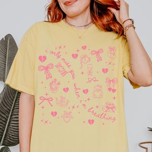 May include: Yellow t-shirt with a pink graphic design featuring hearts, bows, and the text "i only miss you when i'm breathing". The shirt has short sleeves and a relaxed fit.