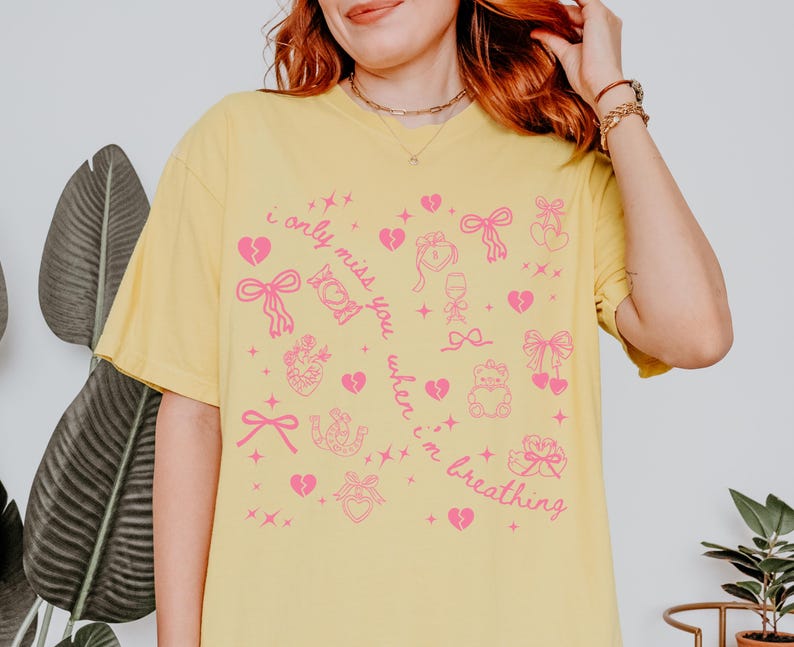 May include: Yellow t-shirt with a pink graphic design featuring hearts, bows, and the text "i only miss you when i'm breathing". The shirt has short sleeves and a relaxed fit.