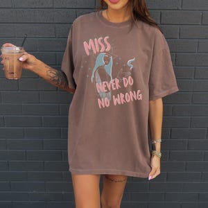 May include: Brown t-shirt with the text "MISS NEVER DO NO WRONG" in pink. The graphic features a woman with blue hair. The shirt is a relaxed fit.