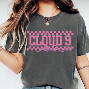 Puede incluir: Camiseta gris oscuro con el texto "CLOUD 9" en rosa, enmarcado por un patr&oacute;n de cuadros rosas y blancos. Un dise&ntilde;o de cinta rosa est&aacute; debajo del texto. La camiseta tiene mangas cortas y cuello redondo.