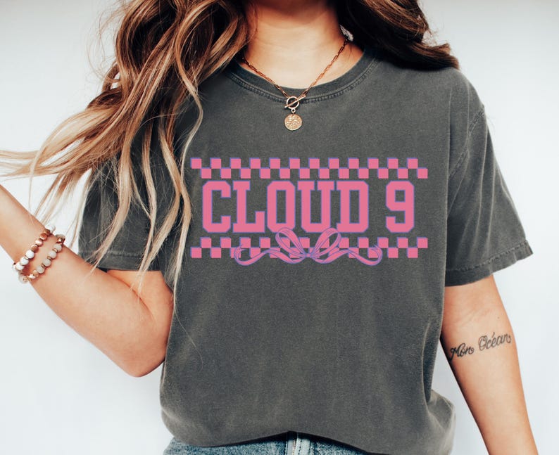 Puede incluir: Camiseta gris oscuro con el texto "CLOUD 9" en rosa, enmarcado por un patr&oacute;n de cuadros rosas y blancos. Un dise&ntilde;o de cinta rosa est&aacute; debajo del texto. La camiseta tiene mangas cortas y cuello redondo.