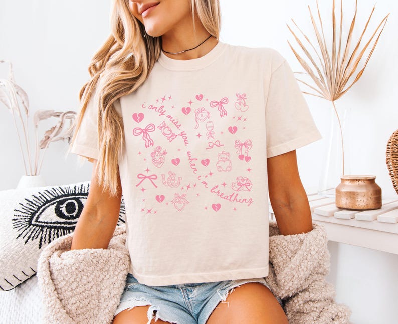 Pode incluir: Camiseta creme com um design gr&aacute;fico rosa. O design inclui cora&ccedil;&otilde;es, la&ccedil;os e o texto "I only miss you when I'm breathing". A camisa &eacute; de estilo casual.
