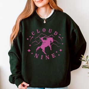K&ouml;nnte beinhalten: Dunkelgr&uuml;nes Sweatshirt mit dem Schriftzug "CLOUD NINE" in Pink und einer pinkfarbenen Grafik einer Person, die auf einem Pferd reitet. Das Sweatshirt hat einen Rundhalsausschnitt und lange &Auml;rmel.