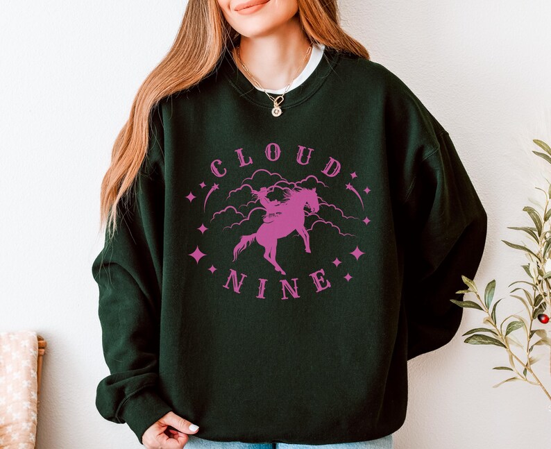 K&ouml;nnte beinhalten: Dunkelgr&uuml;nes Sweatshirt mit dem Schriftzug "CLOUD NINE" in Pink und einer pinkfarbenen Grafik einer Person, die auf einem Pferd reitet. Das Sweatshirt hat einen Rundhalsausschnitt und lange &Auml;rmel.