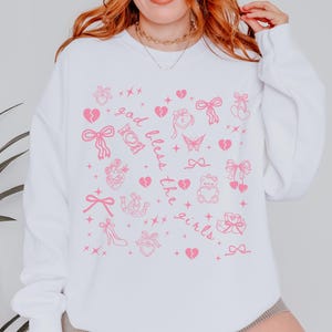 Może przedstawiać: Biała bluza z r&oacute;żowymi grafikami przedstawiającymi kokardy, serca, motyle i napisem "God bless the girls". Wz&oacute;r powtarza się na przodzie bluzy.