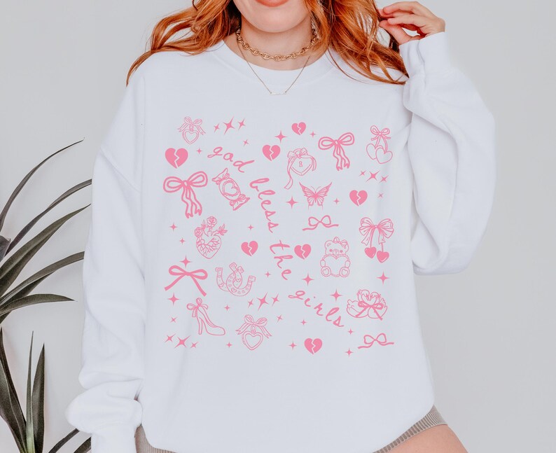 Może przedstawiać: Biała bluza z r&oacute;żowymi grafikami przedstawiającymi kokardy, serca, motyle i napisem "God bless the girls". Wz&oacute;r powtarza się na przodzie bluzy.