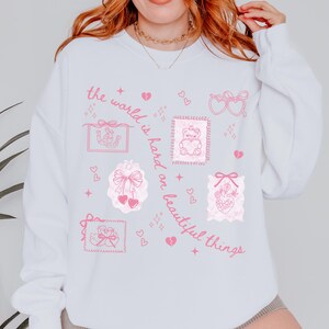 K&ouml;nnte beinhalten: Wei&szlig;es Sweatshirt mit rosa grafischen Illustrationen, darunter Herzen, Schleifen und ein Teddyb&auml;r, der ein Herz h&auml;lt. Der Text "the world is hard on beautiful things" ist in rosa Schrift geschrieben.