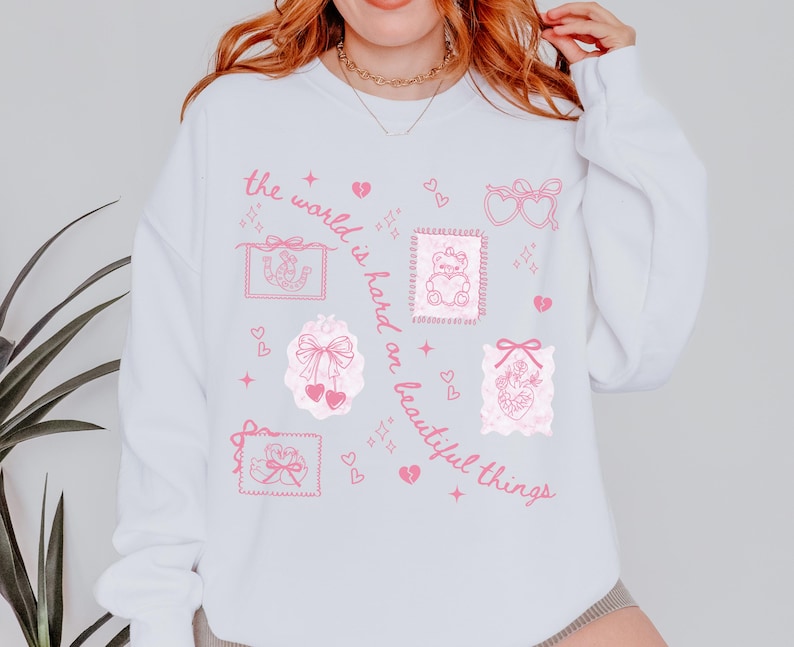 K&ouml;nnte beinhalten: Wei&szlig;es Sweatshirt mit rosa grafischen Illustrationen, darunter Herzen, Schleifen und ein Teddyb&auml;r, der ein Herz h&auml;lt. Der Text "the world is hard on beautiful things" ist in rosa Schrift geschrieben.