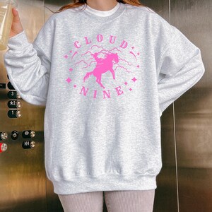 K&ouml;nnte beinhalten: Hellgraues Sweatshirt mit einem rosa Aufdruck, der ein Einhorn, Sterne und den Schriftzug "Cloud Nine" zeigt. Das Sweatshirt hat lange &Auml;rmel und einen Rundhalsausschnitt.