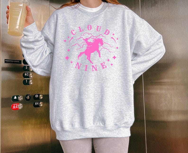 K&ouml;nnte beinhalten: Hellgraues Sweatshirt mit einem rosa Aufdruck, der ein Einhorn, Sterne und den Schriftzug "Cloud Nine" zeigt. Das Sweatshirt hat lange &Auml;rmel und einen Rundhalsausschnitt.