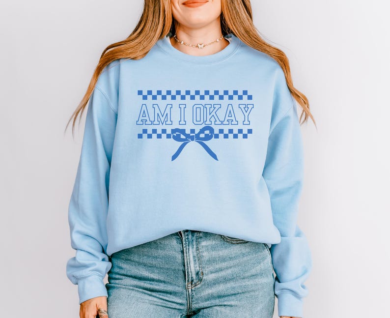May include: Light blue sweatshirt with the text "AM I OKAY" in a blue, checkered design. A blue bow graphic is below the text. The sweatshirt has long sleeves and a crew neck.