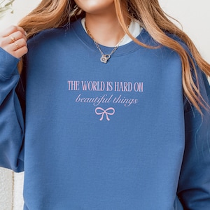 Może przedstawiać: Niebieska bluza z r&oacute;żowym napisem "THE WORLD IS HARD ON beautiful things" i r&oacute;żową grafiką kokardy. Bluza ma długie rękawy i okrągły dekolt.