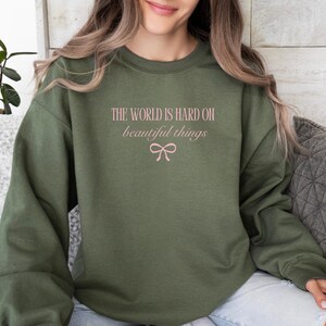 Może przedstawiać: Zielona bluza z napisem "THE WORLD IS HARD ON beautiful things" w kolorze r&oacute;żowym i grafiką kokardy. Bluza z długim rękawem i okrągłym dekoltem to swobodny styl.