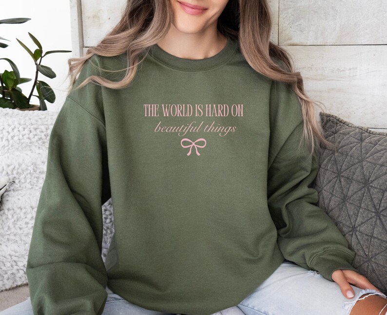 Może przedstawiać: Zielona bluza z napisem "THE WORLD IS HARD ON beautiful things" w kolorze r&oacute;żowym i grafiką kokardy. Bluza z długim rękawem i okrągłym dekoltem to swobodny styl.
