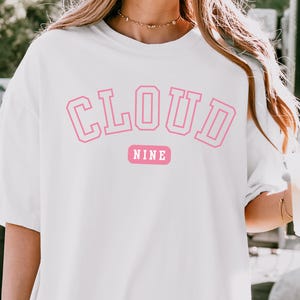 May include: White t-shirt with the words "CLOUD NINE" in pink, outlined block letters. Below the word "CLOUD" is the word "NINE" in a pink rectangle. The shirt is a casual, oversized style.