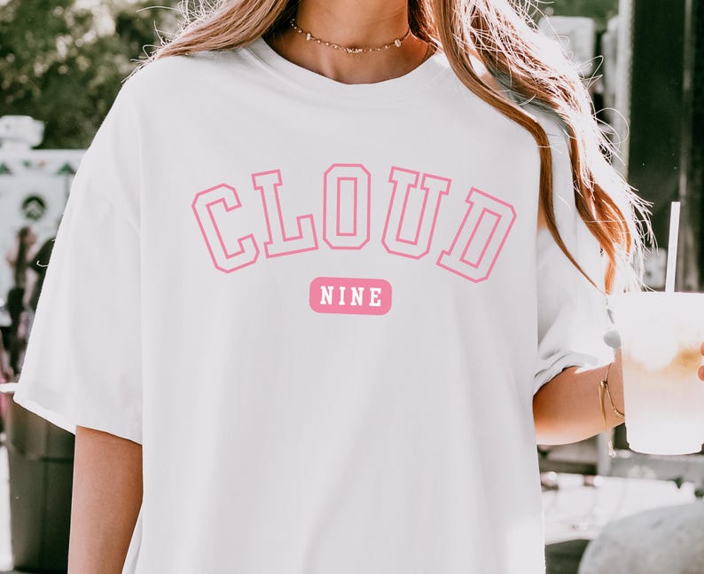 May include: White t-shirt with the words "CLOUD NINE" in pink, outlined block letters. Below the word "CLOUD" is the word "NINE" in a pink rectangle. The shirt is a casual, oversized style.