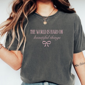 Puede incluir: Camiseta gris oscuro con el texto rosa "THE WORLD IS HARD ON beautiful things" y un lazo rosa. Una camiseta informal y c&oacute;moda para el uso diario.