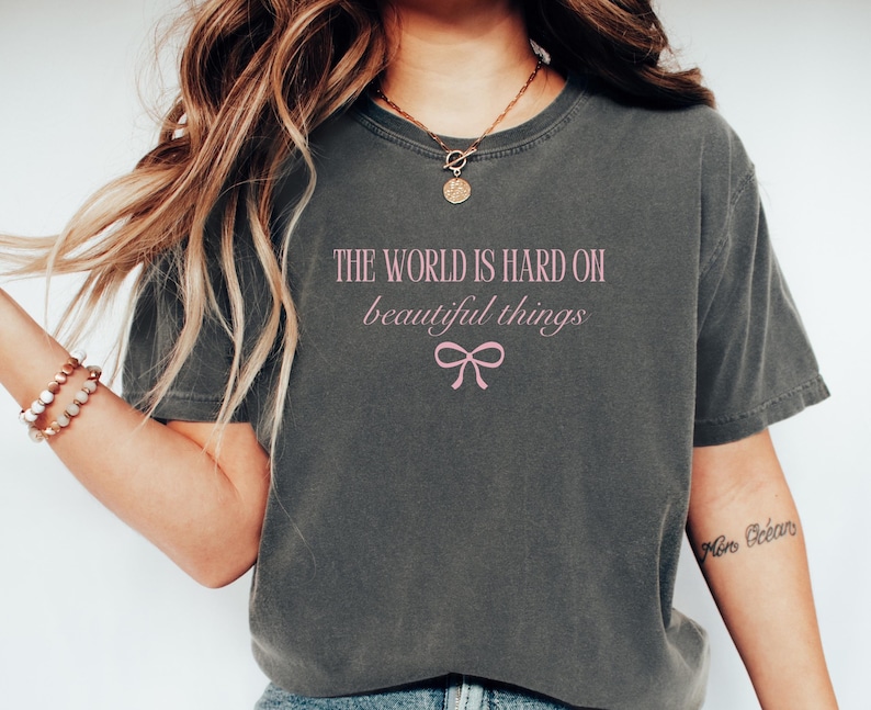 Puede incluir: Camiseta gris oscuro con el texto rosa "THE WORLD IS HARD ON beautiful things" y un lazo rosa. Una camiseta informal y c&oacute;moda para el uso diario.