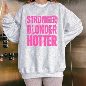 Puede incluir: Una sudadera gris claro con las palabras "STRONGER BLONDER HOTTER" en letras rosas brillantes. El texto est&aacute; apilado verticalmente. La sudadera tiene mangas largas y cuello redondo.