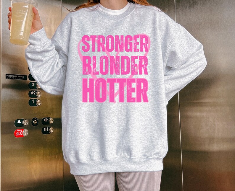 Puede incluir: Una sudadera gris claro con las palabras "STRONGER BLONDER HOTTER" en letras rosas brillantes. El texto est&aacute; apilado verticalmente. La sudadera tiene mangas largas y cuello redondo.