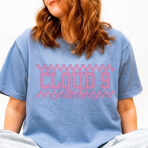 Puede incluir: Camiseta azul claro con la inscripci&oacute;n rosa "CLOUD 9" sobre un dise&ntilde;o decorativo. El texto est&aacute; sobre un patr&oacute;n de cuadros rosas y blancos. La camiseta tiene un corte casual y holgado.