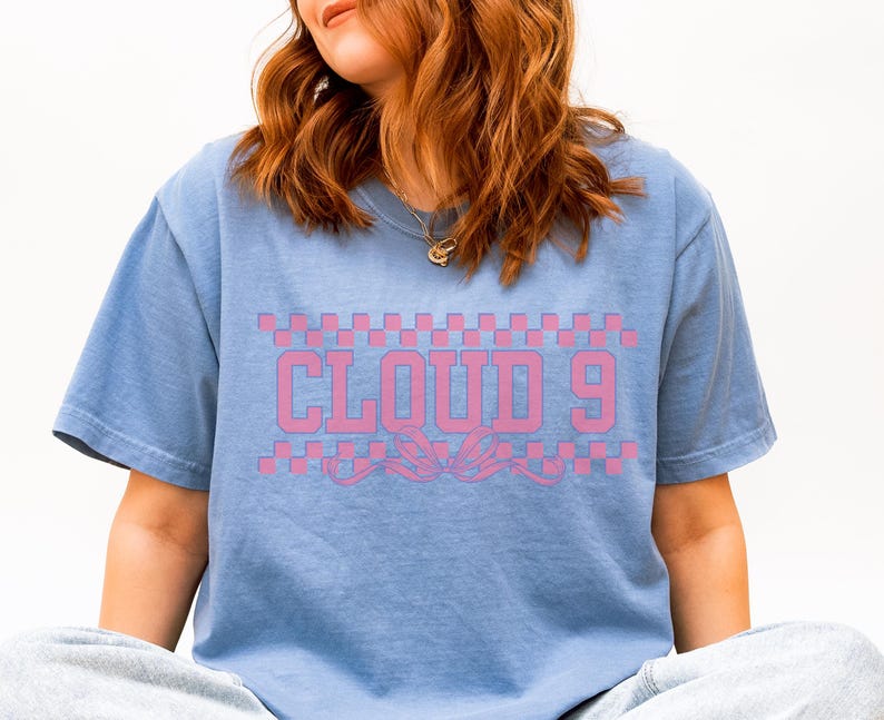 Puede incluir: Camiseta azul claro con la inscripci&oacute;n rosa "CLOUD 9" sobre un dise&ntilde;o decorativo. El texto est&aacute; sobre un patr&oacute;n de cuadros rosas y blancos. La camiseta tiene un corte casual y holgado.