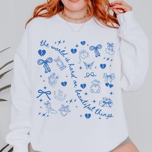 May include: White sweatshirt with blue illustrations of bows, hearts, and other symbols. The text "the world is hard on beautiful things" is printed in blue. The sweatshirt is a casual style.