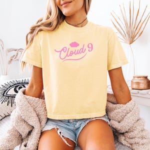 May include: A light yellow t-shirt with the text "Cloud 9" in pink script and a pink cloud graphic. The shirt is short-sleeved and appears to be made of a soft material. The model is wearing denim shorts and a beige sweater.