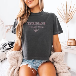 May include: Dark gray t-shirt with the text "THE WORLD IS HARD ON beautiful things" in pink script, and a pink bow graphic. The shirt is worn with denim shorts and a cream-colored sweater.