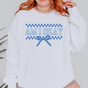 May include: White sweatshirt with the text "AM I OKAY" in blue, with a blue bow graphic. The text is framed by a blue and white checkered pattern. The sweatshirt has long sleeves.