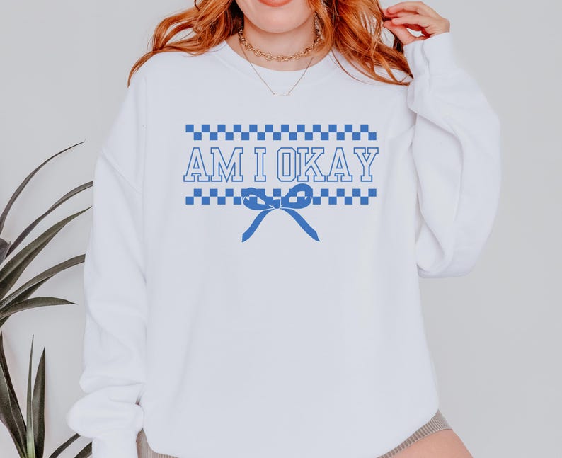 May include: White sweatshirt with the text "AM I OKAY" in blue, with a blue bow graphic. The text is framed by a blue and white checkered pattern. The sweatshirt has long sleeves.