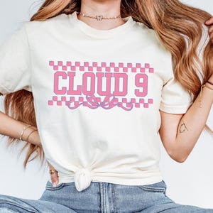 Puede incluir: Camiseta color crema con el texto "CLOUD 9" en letras rosas, enmarcado por un patr&oacute;n de cuadros. Un dise&ntilde;o de cinta decorativa est&aacute; debajo del texto. Es una prenda casual para el uso diario.