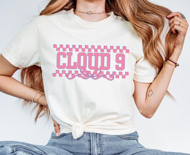 Puede incluir: Camiseta color crema con el texto "CLOUD 9" en letras rosas, enmarcado por un patr&oacute;n de cuadros. Un dise&ntilde;o de cinta decorativa est&aacute; debajo del texto. Es una prenda casual para el uso diario.