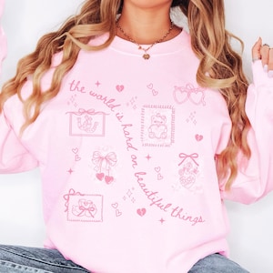 K&ouml;nnte beinhalten: Hellrosa Sweatshirt mit dem Text "the world is hard on beautiful things" und verschiedenen rosa Grafiken, darunter Herzen, Schleifen und ein Teddyb&auml;r. Das lang&auml;rmlige Oberteil ist ein l&auml;ssiger Stil.